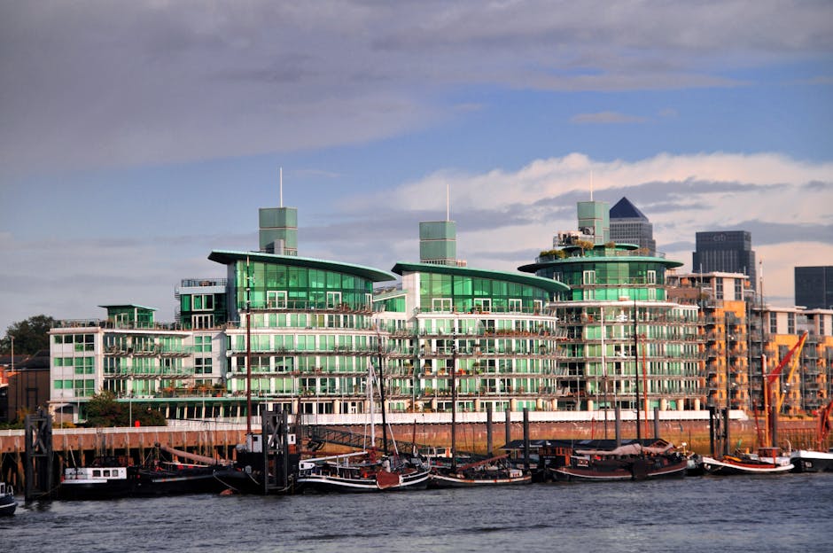 A modern multi-storey building with a distinctive architectural design featuring curved green glass facades and rooftop structures, situated along a waterfront. In the foreground, a wooden dock is visible, with several moored boats and yachts, some with covers, tied to the port, indicating a tranquil marina setting. The building appears to be part of a residential or commercial complex, with numerous balconies and large windows. Construction equipment and scaffolding can be seen on the right side, suggesting ongoing development or renovations. The overall scene is well-lit with natural daylight, showcasing the contemporary style of the building in the context of a busy, vibrant riverside environment. Chelsea Man and Van may be involved in house or office removals in this area, supporting clients with furniture transport and packing during their relocation process.