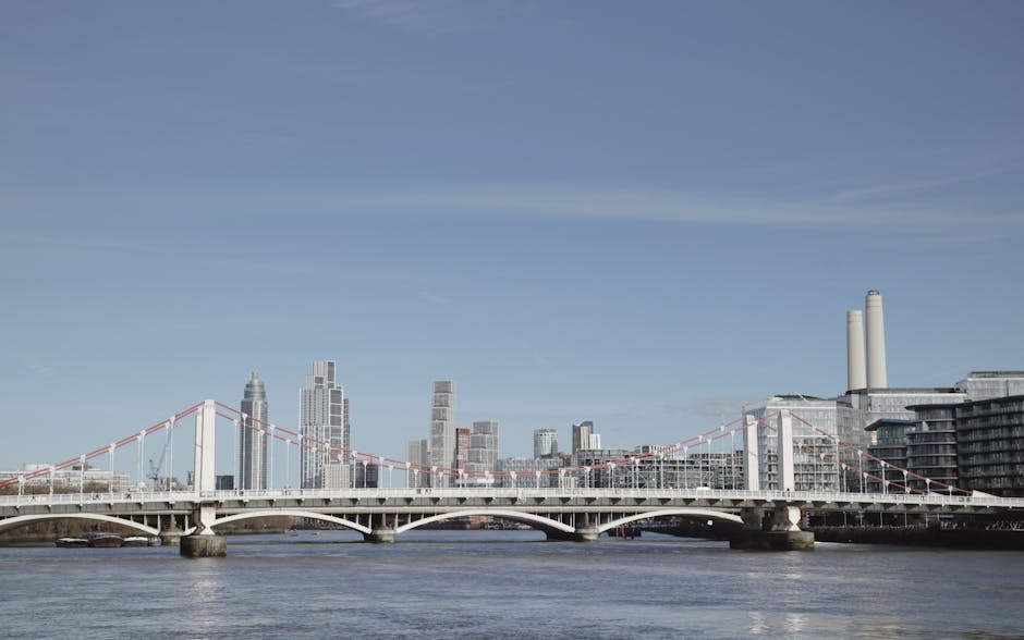 A wide view of the Chelsea Bridge spanning a river in central London, with a modern city skyline featuring tall office buildings and residential structures in the background. The bridge's white suspension cables are visible against a clear blue sky, and the river below reflects the surroundings. On the right side of the image, part of a building with large windows and rooftop terraces can be seen, indicating urban development. This scene captures the urban environment typical of areas where house removals and furniture transport services, such as those provided by Chelsea Man and Van, might operate, especially during home relocations or moving projects near Chelsea Harbour. The setting emphasizes the logistical context of moving and packing within a busy metropolitan area, highlighting the importance of efficient loading processes and transportation across iconic city landmarks.