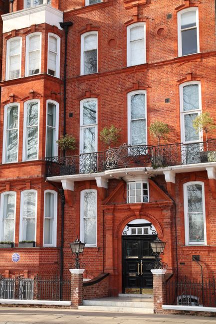 The image shows a multi-storey red brick residential building with tall, white-framed sash windows, some with small decorative arches, and a black front door with a semi-circular fanlight above it. In front of the building, there is a paved pavement with a black iron fence and gate, supported by brick pillars with white caps. Two traditional black lampposts are positioned on either side of the entrance steps, which lead to the doorway. The building features a small balcony with an ornate black metal railing on the second floor, and potted plants are visible on the balcony. Green trees and shrubs are present near the entrance, adding greenery to the scene, with natural daylight illuminating the facade. This setting captures the exterior of a classic London home potentially being prepared for a house removal or furniture transport, as indicated by the context of Chelsea Man and Van and moving services.