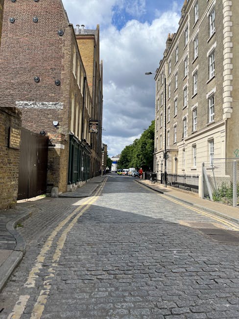 A narrow cobbled street in Chelsea, lined with old brick and stone buildings on both sides, with a mix of residential and commercial properties. On the left, there are small ground-floor entrances with dark green and brown doors, and above some are small windows with limited visibility. The right side features a larger, cream-colored building with multiple windows and decorative architectural details. The street appears to be part of a home relocation or furniture transport process managed by Chelsea Man and Van, as evidenced by a partially visible vehicle at the far end of the street, and the scene suggests a loading or unloading operation taking place amidst the urban setting. The sky above is partly cloudy, with patches of blue visible through the clouds, and overall lighting is natural, indicating daytime. The scene captures the typical tight access challenges encountered in house removals within this historic London neighbourhood.
