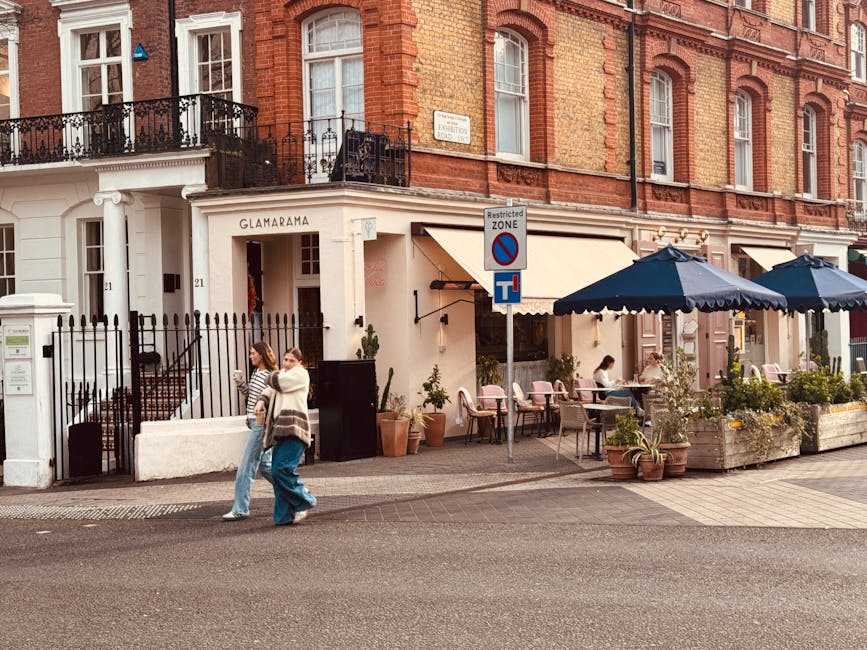 A street scene outside a boutique-style café with outdoor seating under large navy blue umbrellas, positioned on a paved sidewalk in front of a red brick building with ornate iron balconies and white window frames. Potted plants and flowers decorate the café's exterior, and a woman and a man are walking past, both dressed casually. A street sign indicating a restricted zone and parking regulations is mounted on a pole near the sidewalk. The scene is well-lit with natural daylight, suggesting a calm, typical day in Chelsea, with potential relevance to local house removals and furniture transport, especially within busy urban environments like Chelsea for moving antiques or household items, supported subtly by Chelsea Man and Van's services.