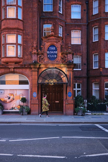 A person wearing a floral dress walking past the entrance of a red-brick building labeled 'The Sloane Club' in Chelsea, with a large wooden door beneath a curved glass canopy. The building features multiple sash windows with white frames and a decorative stone arch surrounding the sign. On the sidewalk in front of the entrance, there are potted plants and small bushes, with the street in the foreground showing a white dashed line and a metal drainage cover. This scene captures the exterior of a residential or private club building, relevant to home relocation and travel logistics, as seen in professional removals and furniture transport activities by Chelsea Man and Van.