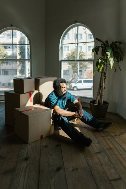 A young man with curly hair and medium skin tone sitting on a wooden floor inside a room with large arched windows, surrounded by several cardboard boxes sealed with red tape, some of which are stacked to the side. The boxes appear to be packed for a home relocation, with the man taking a break during packing or moving activities. Natural daylight streams in through the windows, illuminating the space and casting soft shadows on the floor. In the corner beside the man, there is a potted plant with long, broad leaves, adding a touch of greenery to the room. This scene reflects the packing process typical of furniture transport and house removals, which Chelsea Man and Van provides as part of their professional removals services, involving household packing, fragile items handling, and preparing belongings for transportation.