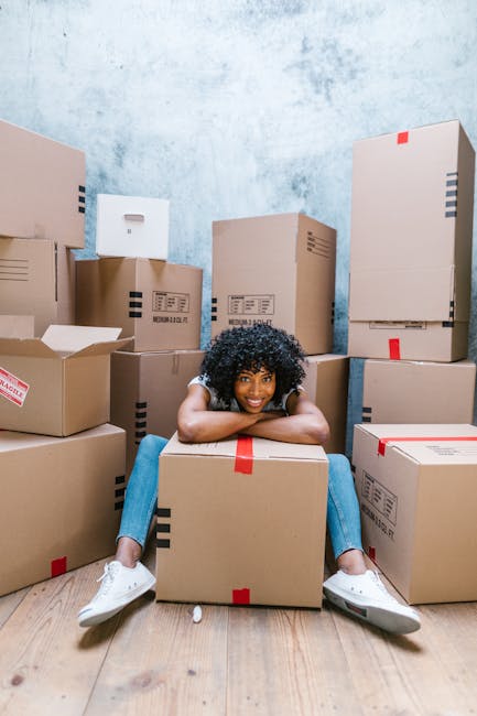 A young woman with curly black hair, wearing a white t-shirt, blue jeans, and white sneakers, sitting on the wooden floor of a room surrounded by numerous cardboard boxes of various sizes, some closed and sealed with red tape, others partially open revealing packing materials. The boxes are stacked along the walls, which have a plain, light-colored textured surface, and the scene appears to depict a home relocation or moving process facilitated by Chelsea Man and Van. The woman is smiling, resting her chin on her arms, and sitting on top of a large box, illustrating a moment during packing or unpacking in a house removal or furniture transport scenario.